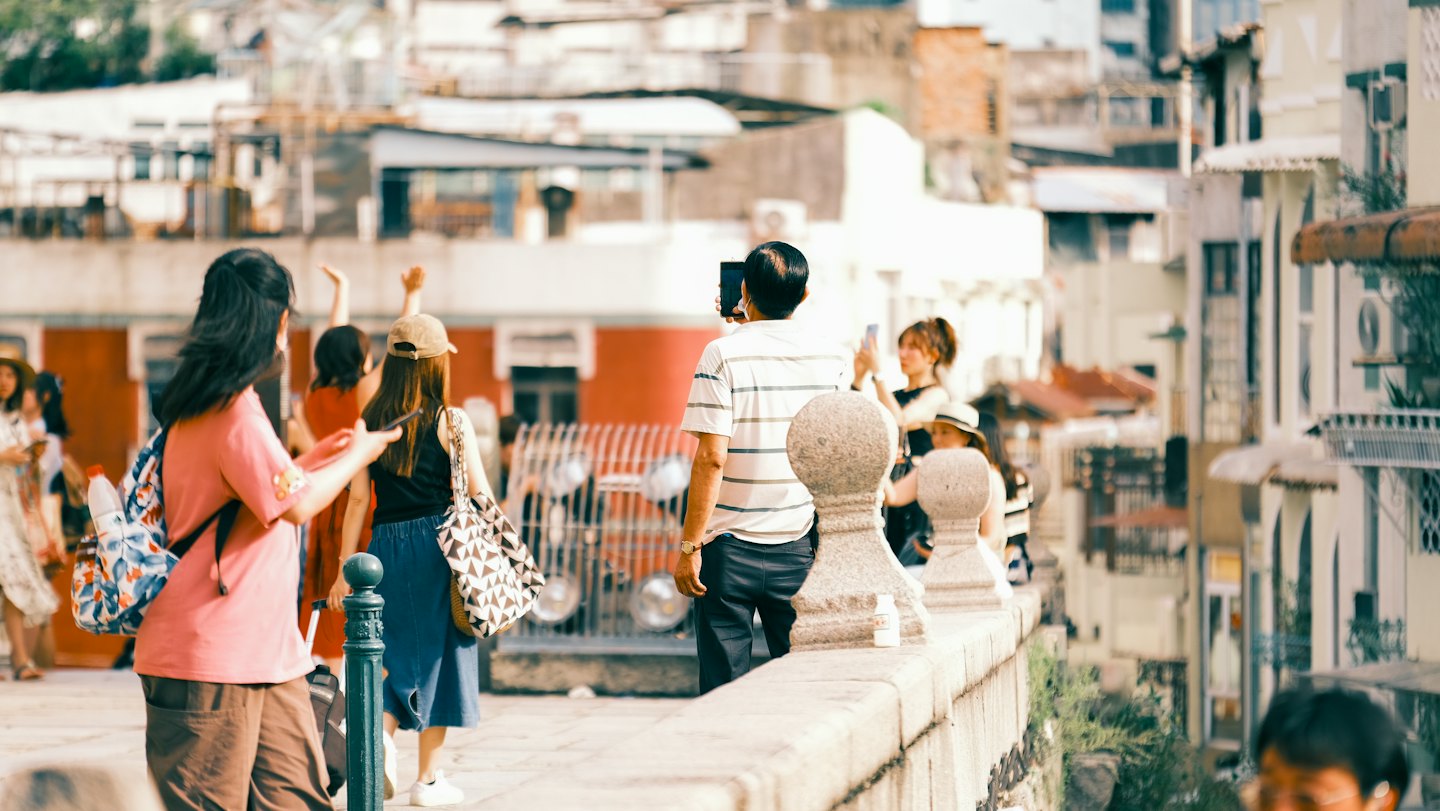 Tourists walk along a stone balustrade while filming with smartphones.
